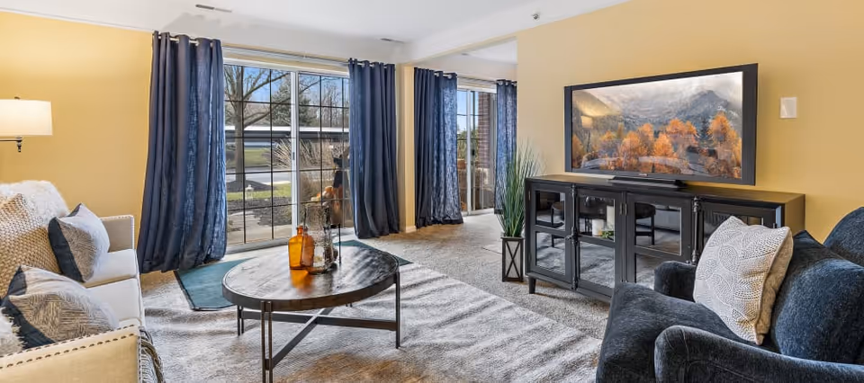 Bright living room with sofas, a round coffee table, TV on a console, and large windows with blue curtains leading to a sliding glass door.