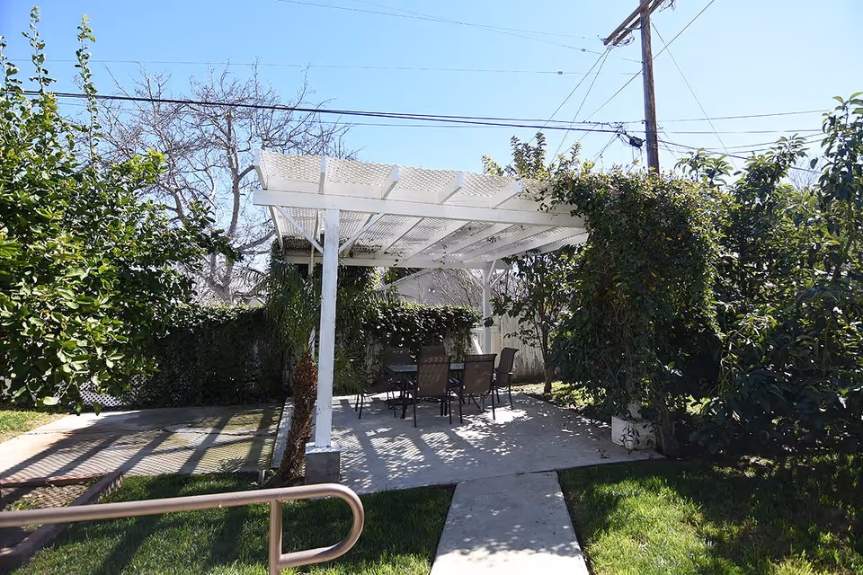 Outdoor patio with a white pergola covering a table and chairs surrounded by trees and shrubs.