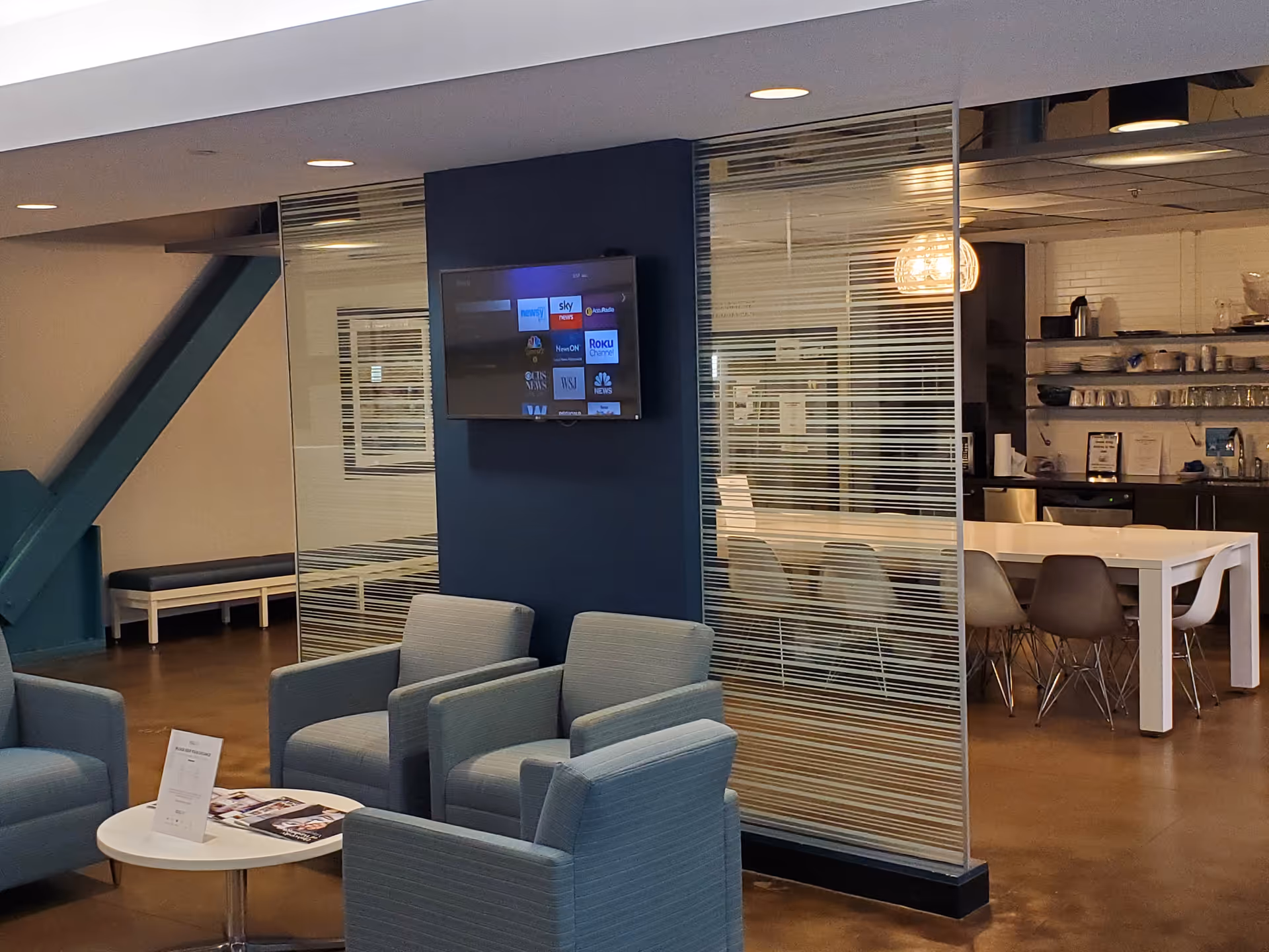 Modern communal lounge with blue seating, glass partitions, and a dining/kitchen area in back.