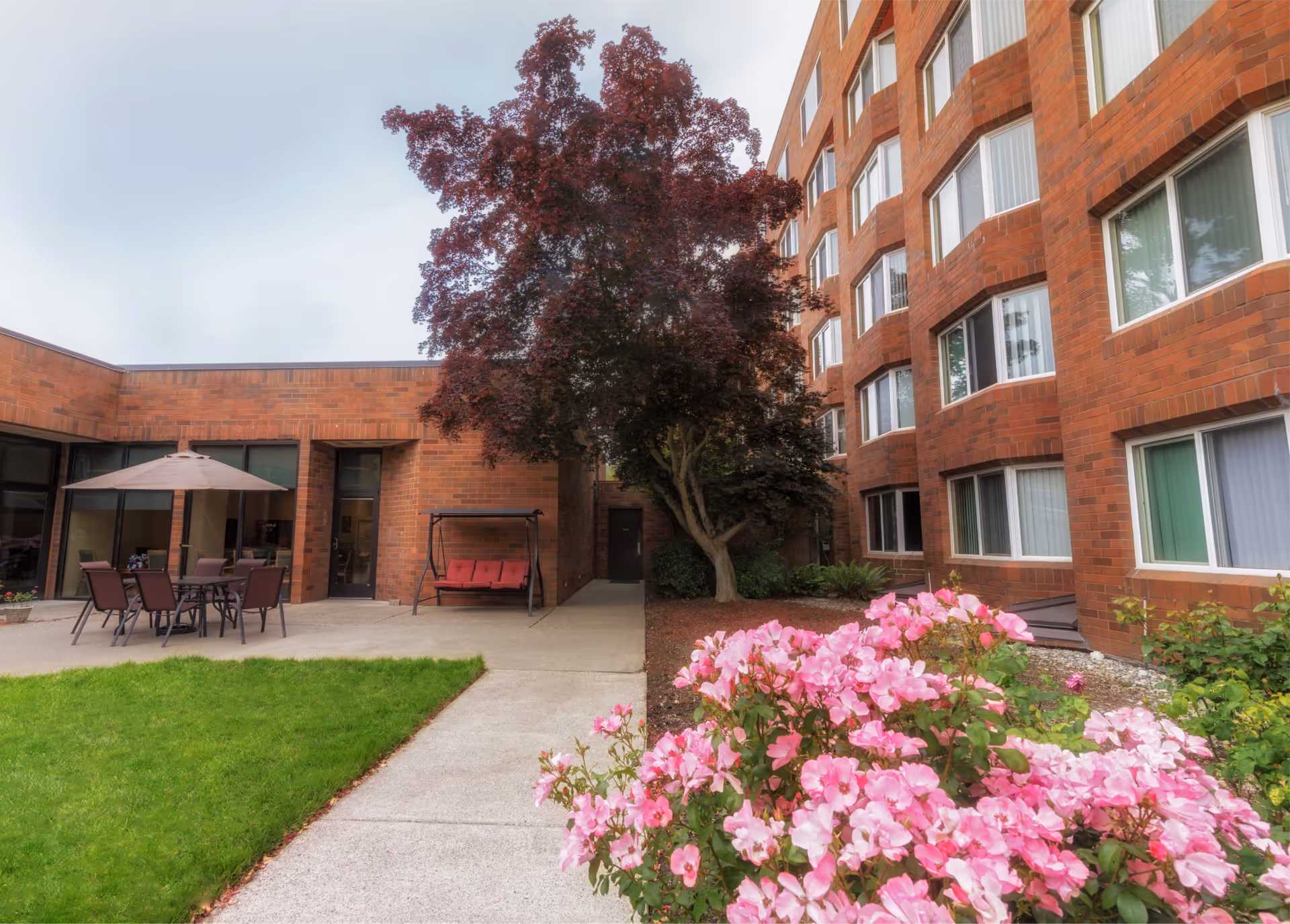 Brick-faced senior living building with a courtyard featuring patio furniture, a swing, a tree, and pink flowers.