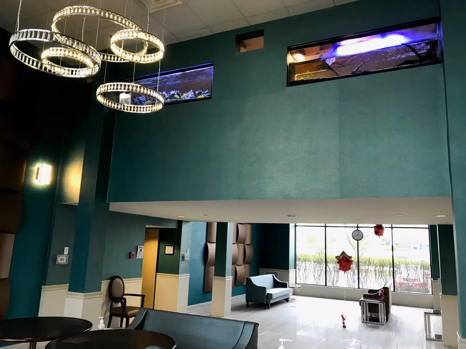 Interior view of a senior living facility lounge area with teal walls, modern circular hanging lights, two built-in aquariums near the ceiling, seating areas with chairs and sofas, large windows letting in natural light, and a clock hanging in front of the window.