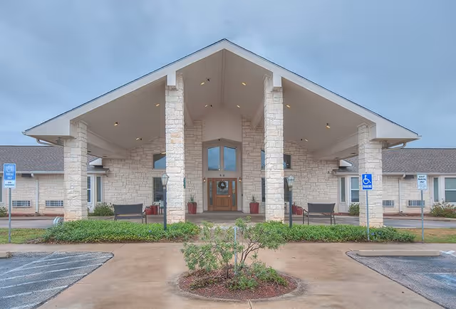 Front exterior view of Heritage Place at Fredericksburg building with stone pillars, a covered entrance, benches, and handicap parking spaces on a cloudy day.