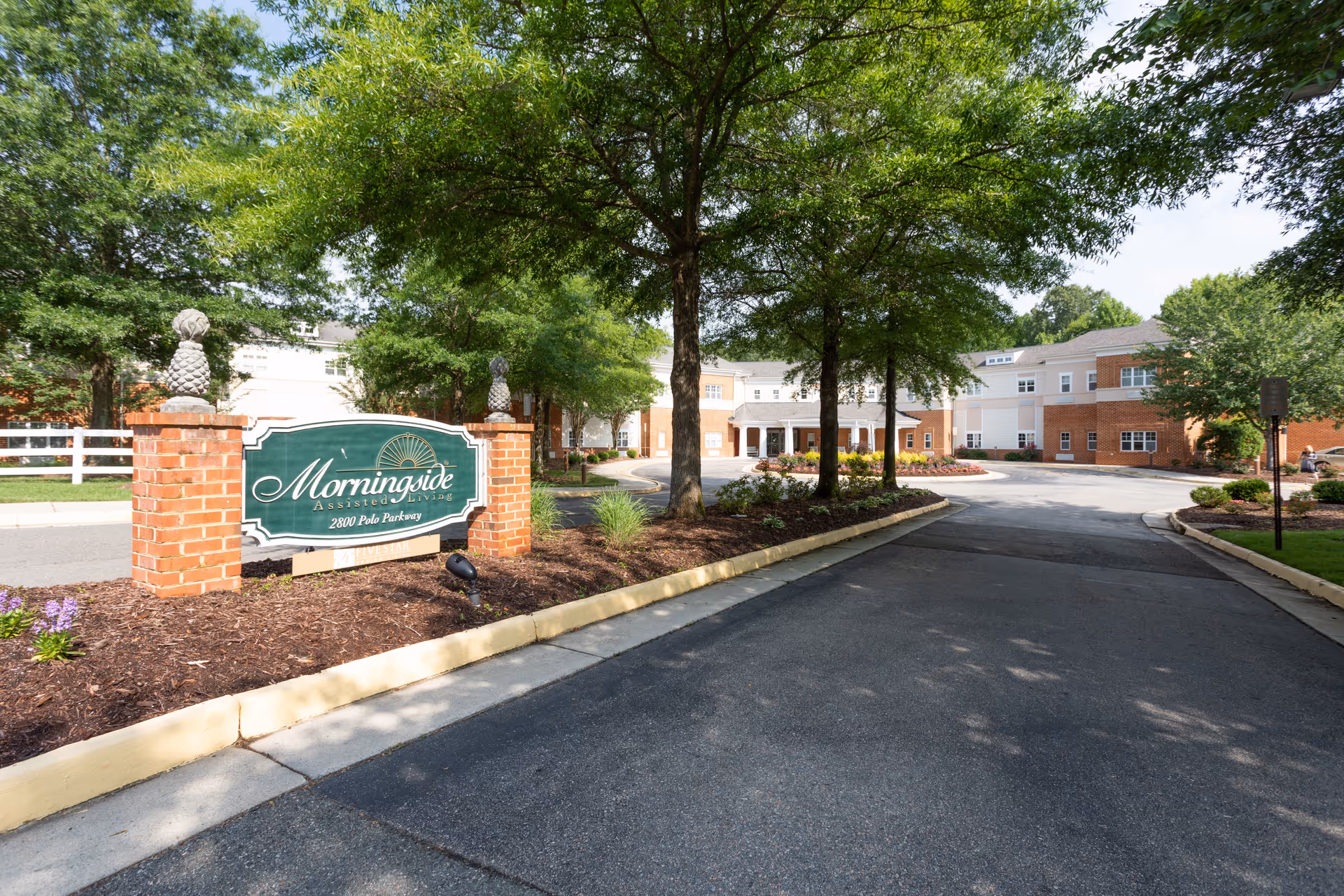 Entrance driveway to Morningside Assisted Living facility with a green sign mounted on brick pillars, surrounded by trees and landscaping, leading to a large two-story building in the background.