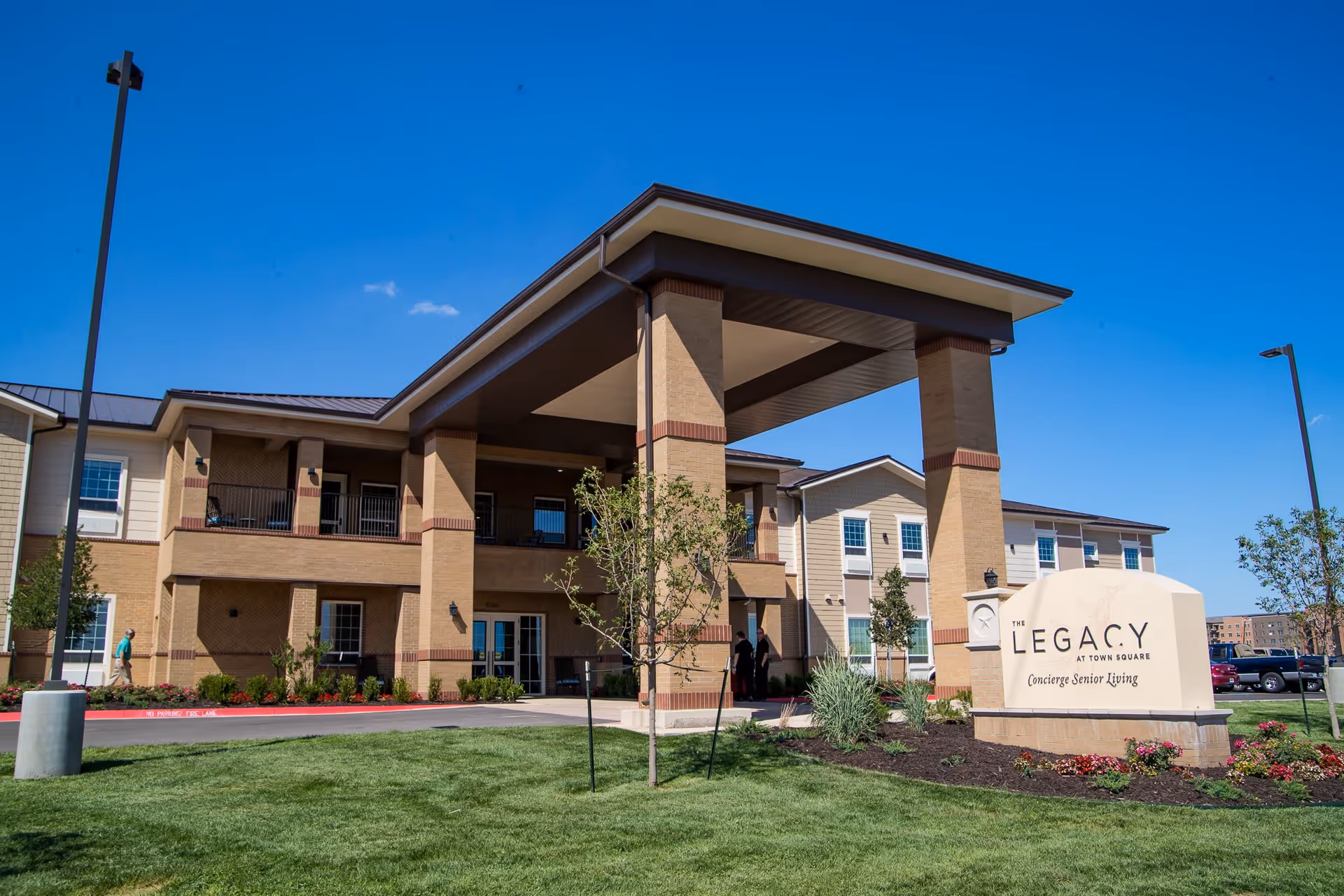 Exterior view of The Legacy at Town Square senior living facility showing a two-story building with a covered entrance, landscaped lawn, and clear blue sky.