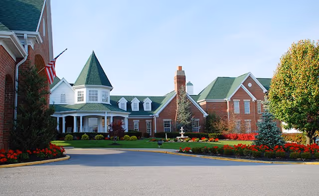 Brick senior living facility with a green-roofed front, turret, flag, and landscaped entrance.