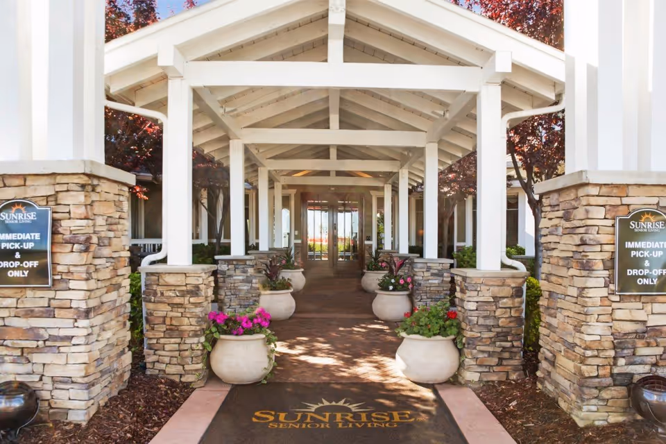 Covered, columned entrance to a Sunrise Senior Living facility framed by stone pillars, potted flowers, and a branded welcome mat.