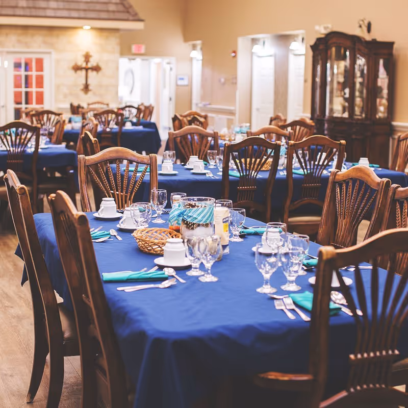 Dining room with multiple wooden tables covered in blue tablecloths, set with glassware, white cups, silverware, and teal napkins. Wooden chairs surround the tables, and a wooden cabinet is visible in the background along with a stone wall and a decorative cross.