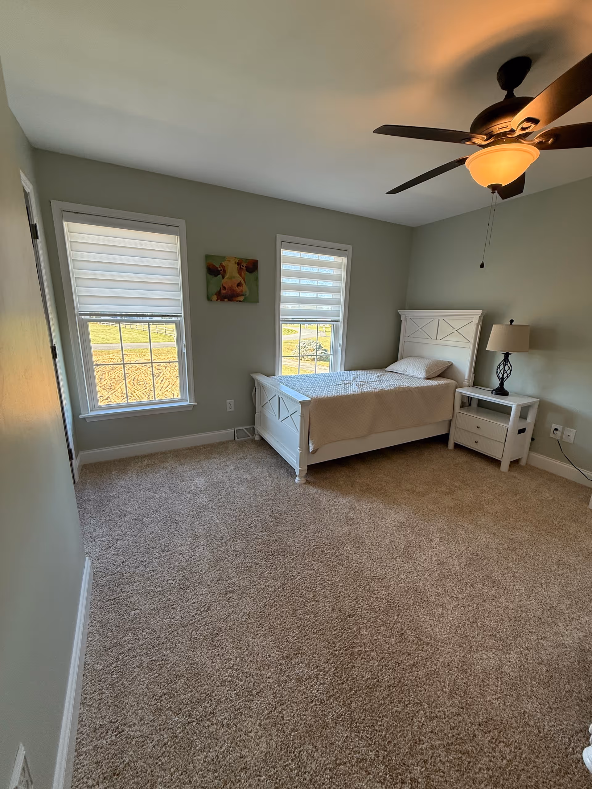 Bright, furnished bedroom with white bed, nightstand, windows, and ceiling fan.