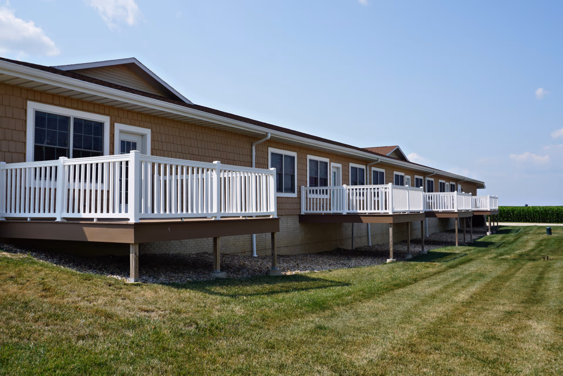 Long single-story assisted living building with a row of white-railed balconies overlooking a grassy lawn under a clear sky.
