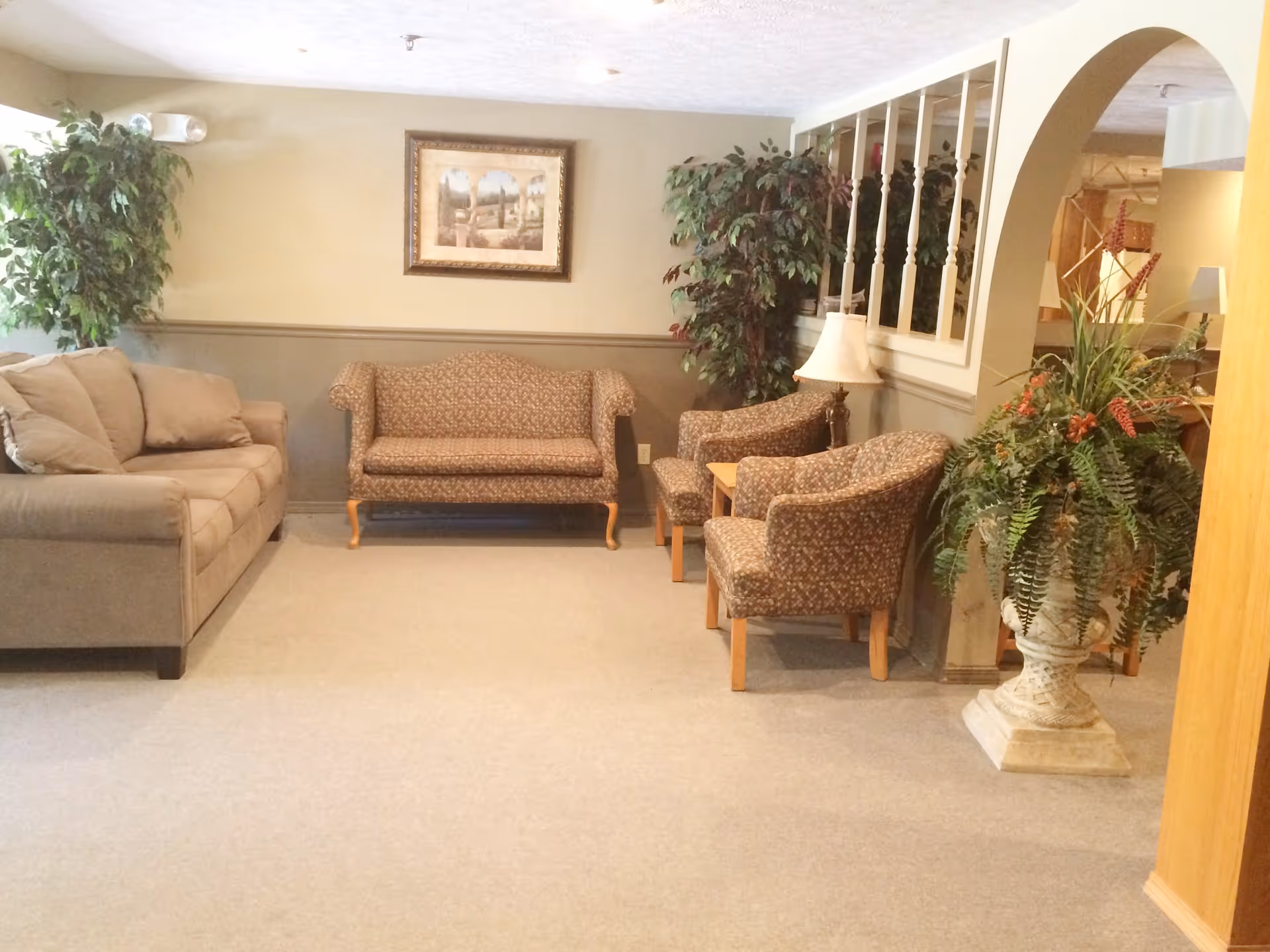 A cozy living room area with a beige sofa, a patterned loveseat, and two matching armchairs. There are two large potted plants, a table lamp, a framed picture on the wall, and a decorative archway with wooden spindles separating the space from another room.