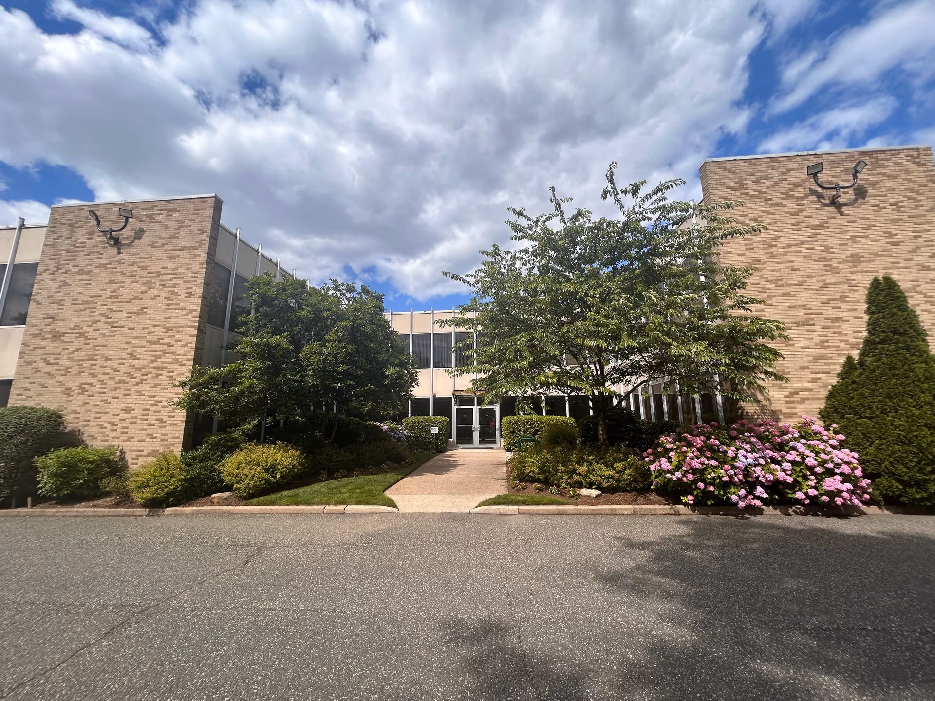 Brick exterior of Life Home Care building with shrubs, trees, and flowering pink flowers at entrance.
