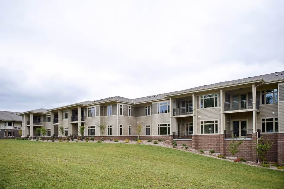 Exterior view of a two-story senior living facility building with beige siding, multiple windows, balconies, and a well-maintained lawn in front under a cloudy sky.