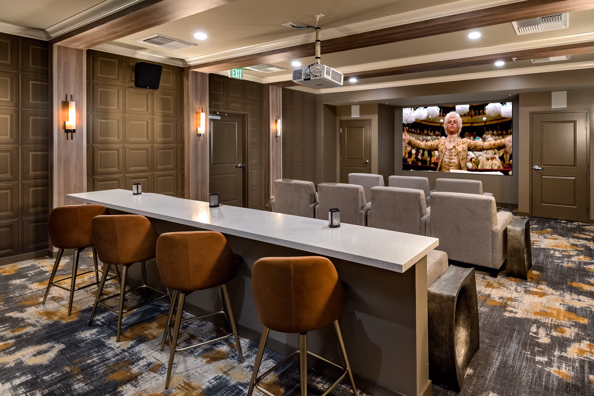 A cozy media room with a large screen showing a historical figure in ornate clothing. The room features a row of four brown leather bar stools at a white countertop, and two rows of beige cushioned chairs facing the screen. The walls are decorated with brown paneling and warm wall sconces, and the ceiling has recessed lighting and a mounted projector.