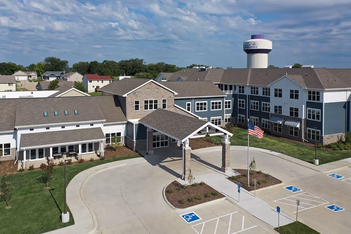 Exterior view of CedarStone Senior Living facility showing a large building with multiple windows, a covered entrance with stone pillars, an American flag on a flagpole, several handicap parking spaces, and a water tower in the background under a partly cloudy sky.