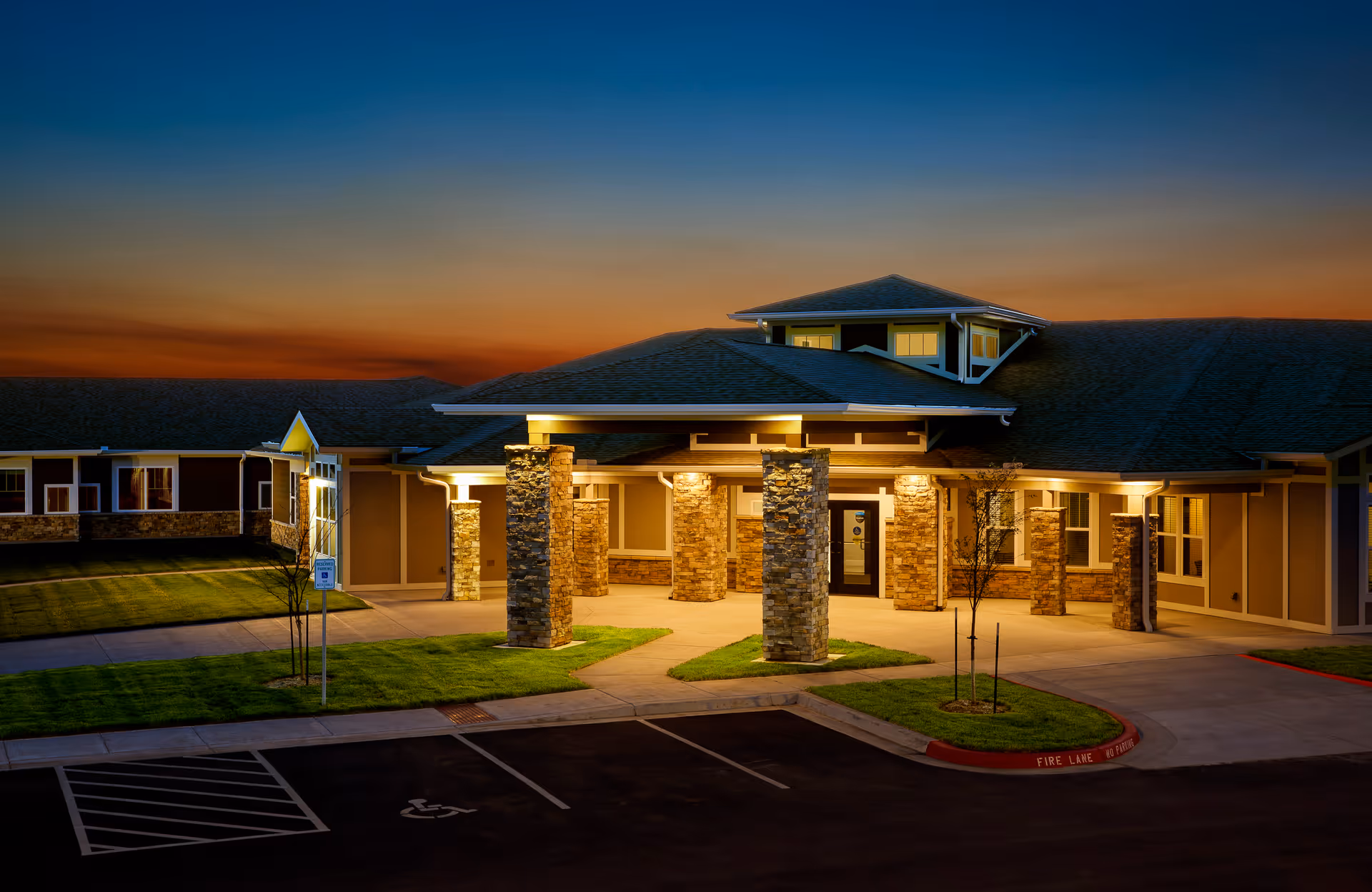 Entrance of a well-lit single-story building with stone columns, landscaped lawn, and a driveway at dusk.
