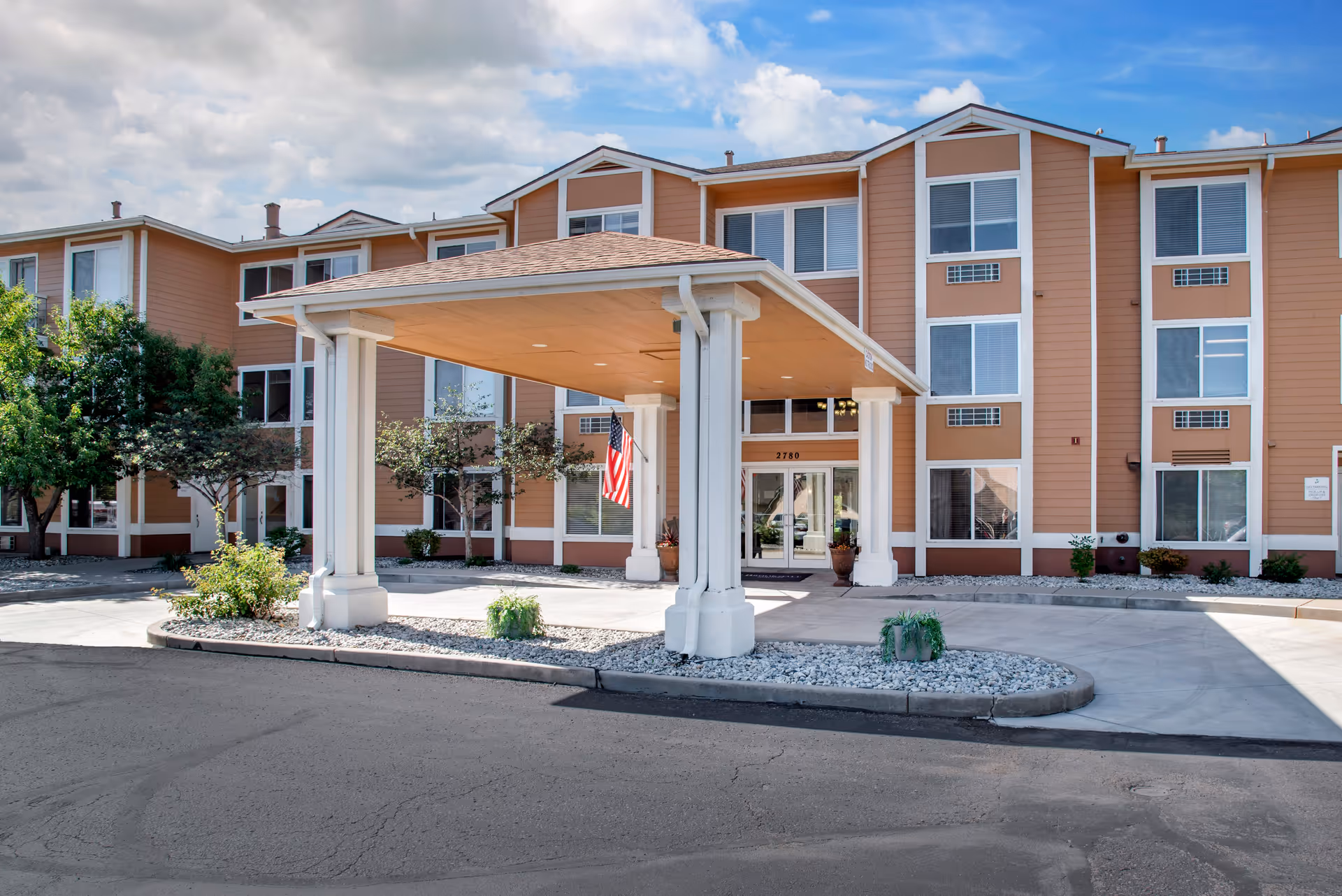 Exterior front view of a three-story senior living facility building with a covered entrance supported by white columns, an American flag hanging near the entrance, and landscaping with small trees and shrubs under a partly cloudy sky.