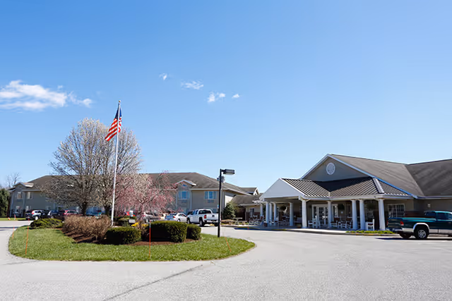 Exterior view of Brookdale Hagerstown senior living facility on a clear day, showing a circular driveway with an American flag on a flagpole in the center, landscaped bushes, and the building entrance with a covered porch and parked vehicles.