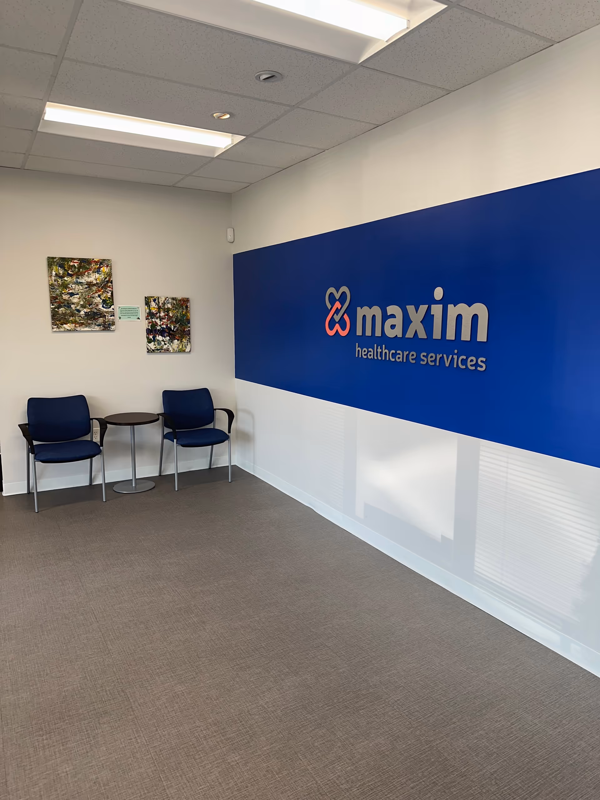 Waiting area with two blue chairs and a round table against a Maxim Healthcare Services wall.
