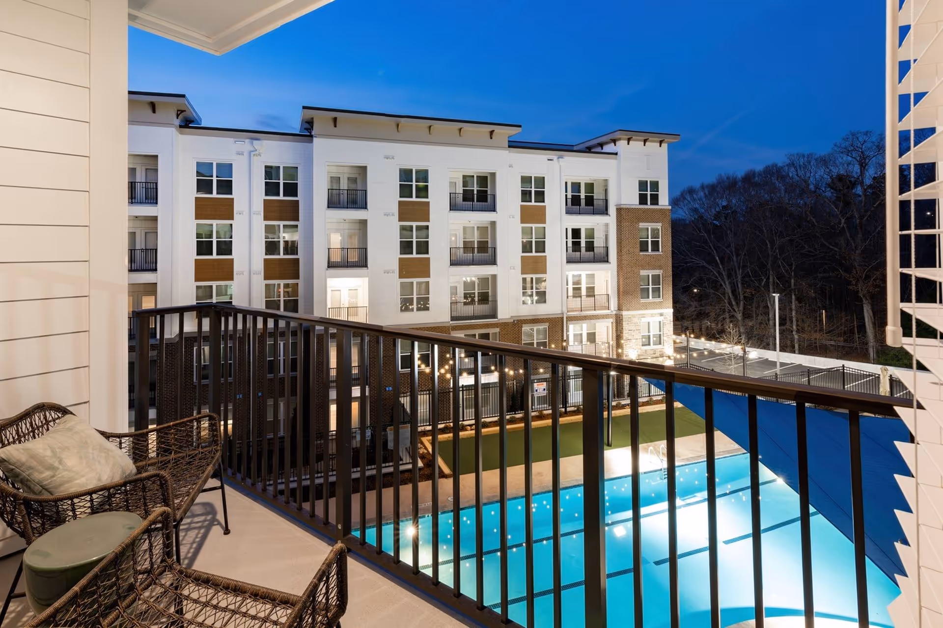 View from a balcony overlooking a swimming pool at night, with a modern multi-story apartment building in the background. The balcony has two wicker chairs with cushions and a small round table. String lights are visible near the pool area.