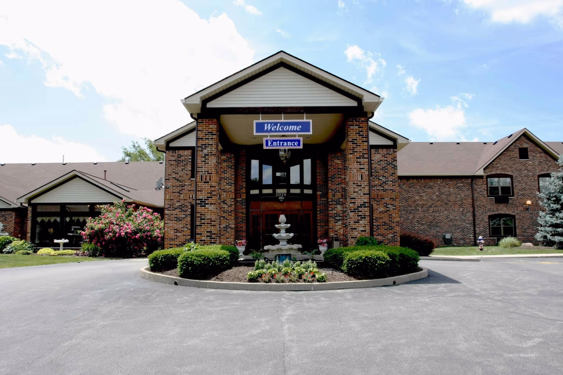 Front exterior view of a brick building with a covered entrance featuring a 'Welcome' and 'Entrance' sign. There is a decorative fountain and landscaped bushes and flowers in front of the entrance under a partly cloudy sky.