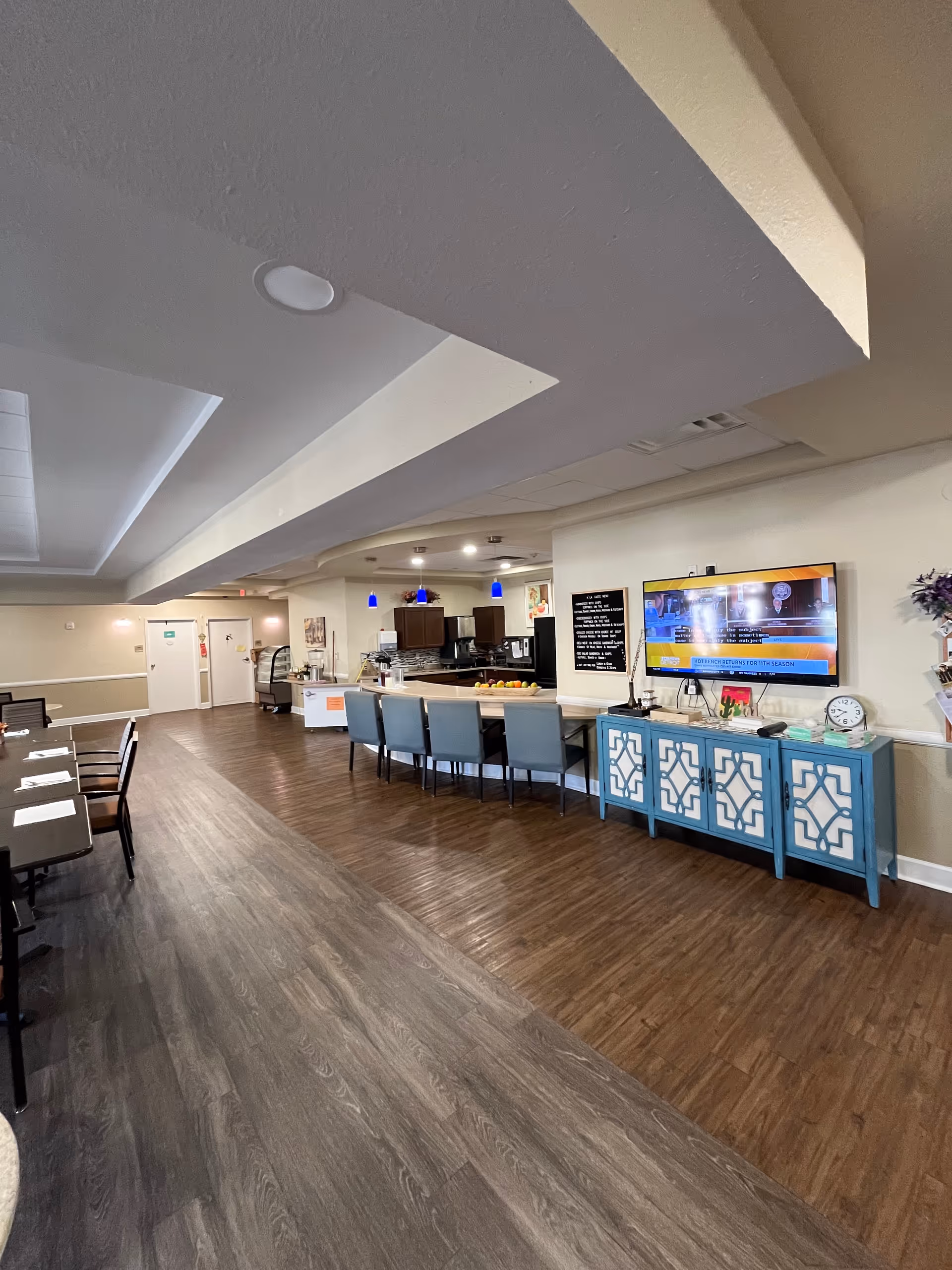Interior senior living dining area with a long counter, teal cabinets, and wall TV.