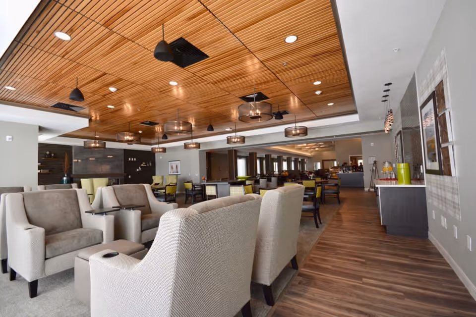 A spacious and modern common area in a senior living facility featuring comfortable armchairs and small tables in the foreground, with dining tables and chairs arranged in the background. The ceiling has wooden paneling with multiple pendant lights, and the floor is a combination of carpet and wood. The walls are decorated with framed artwork and there is a counter area with beverage dispensers on the right side.