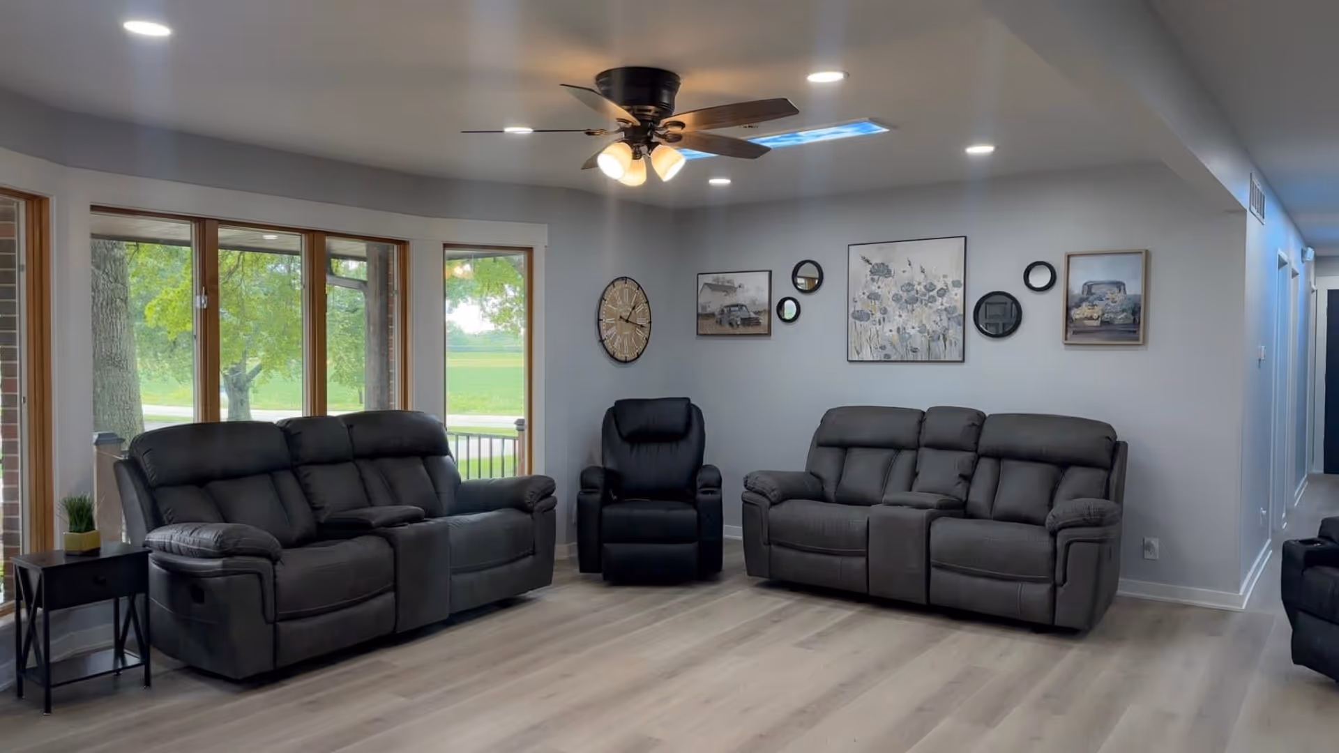 Bright living room with gray recliners, wall art, ceiling fan, and large windows.