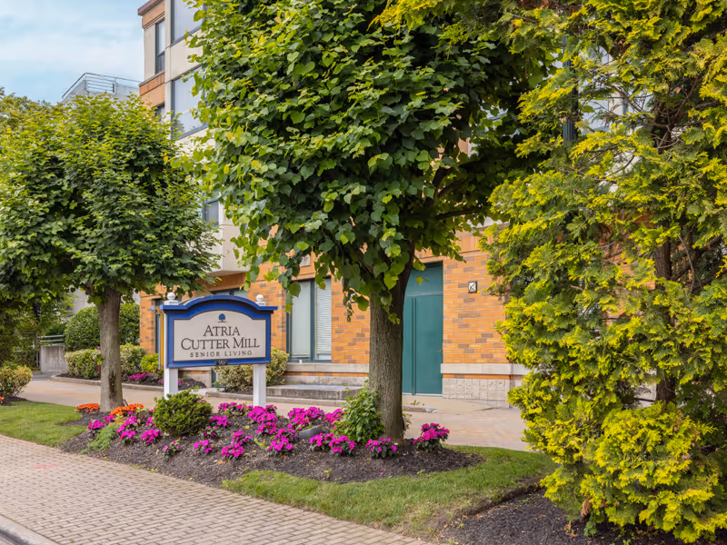 Exterior view of Atria Cutter Mill with a sign, trees, and blooming flowers.