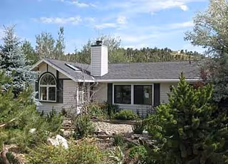 Single-story house with a chimney surrounded by trees, shrubs, and a rocky front garden under a blue sky.