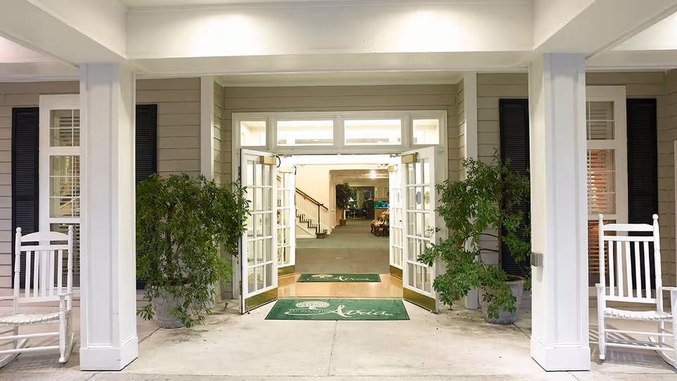 Entrance to a senior living facility with open double glass doors, two large potted plants on either side, white rocking chairs, and a covered porch area.