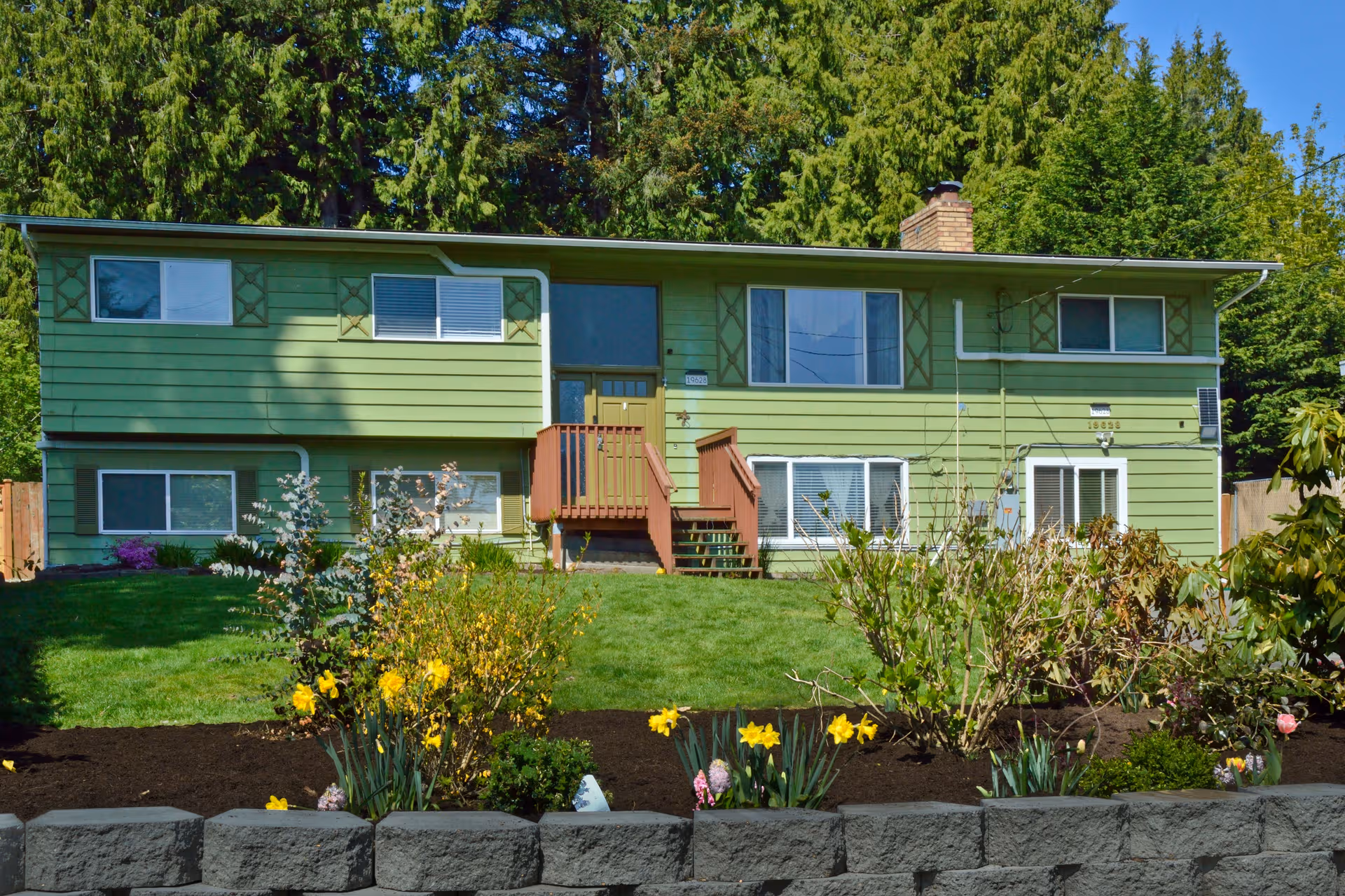 Green two-story building with front steps, lawn, and blooming flower beds.