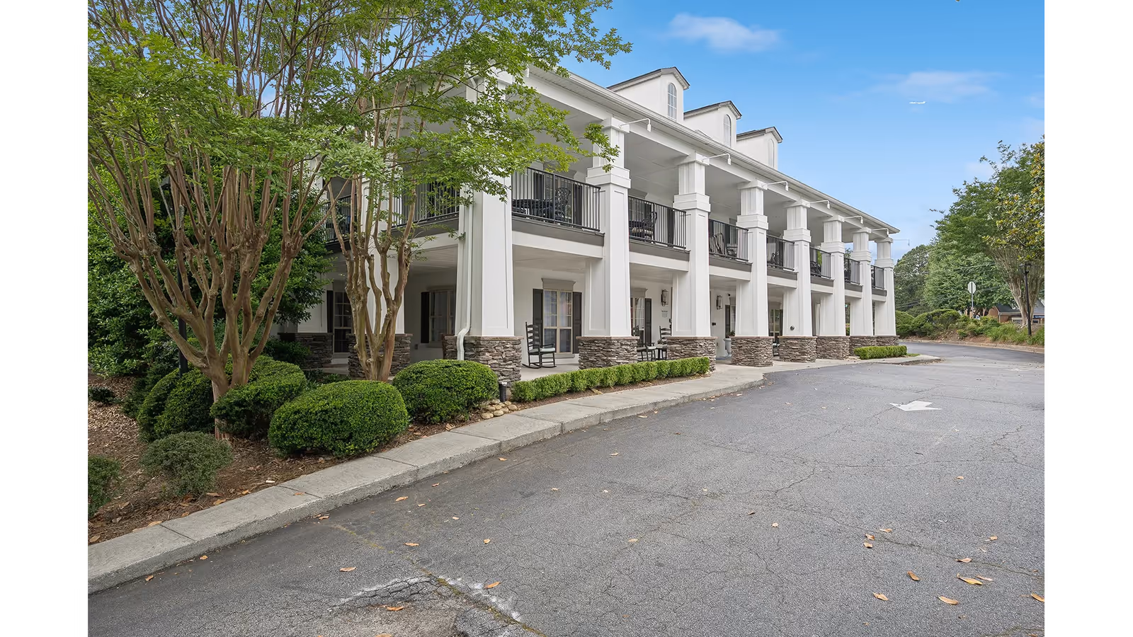 Exterior view of a two-story white columned residential building with balconies, landscaping, and a driveway.
