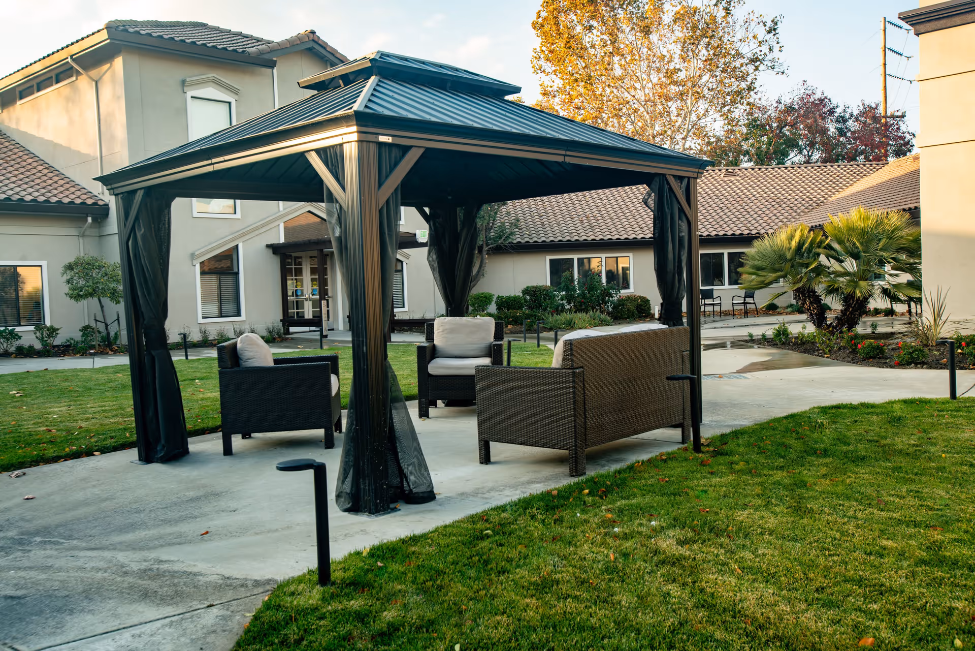 Outdoor seating area with a black metal gazebo covering cushioned wicker chairs and a sofa, situated on a concrete patio surrounded by green grass and landscaping, with beige buildings and trees in the background.