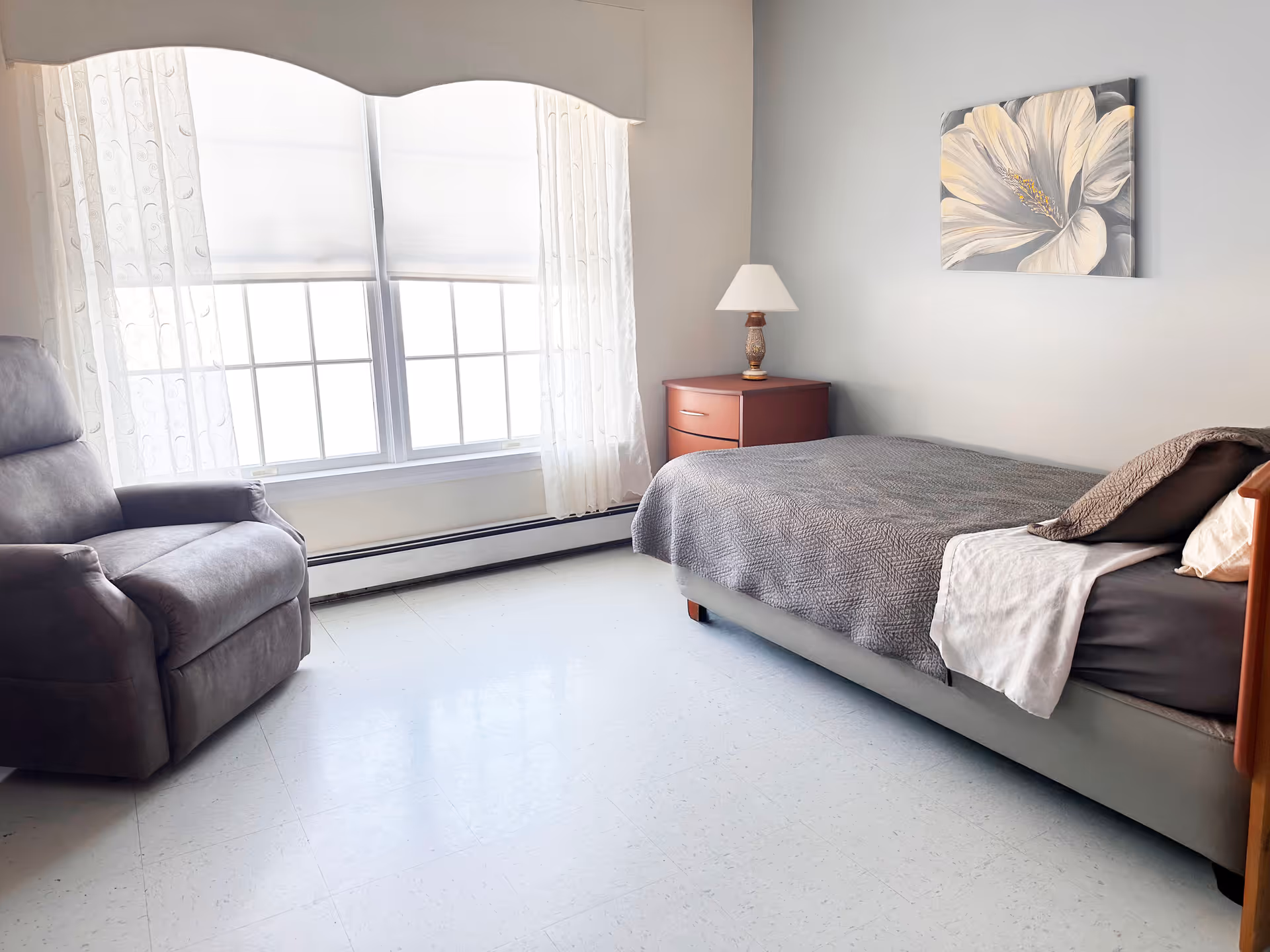 A bedroom featuring a bed, bedside table with lamp, and a recliner by a large window.