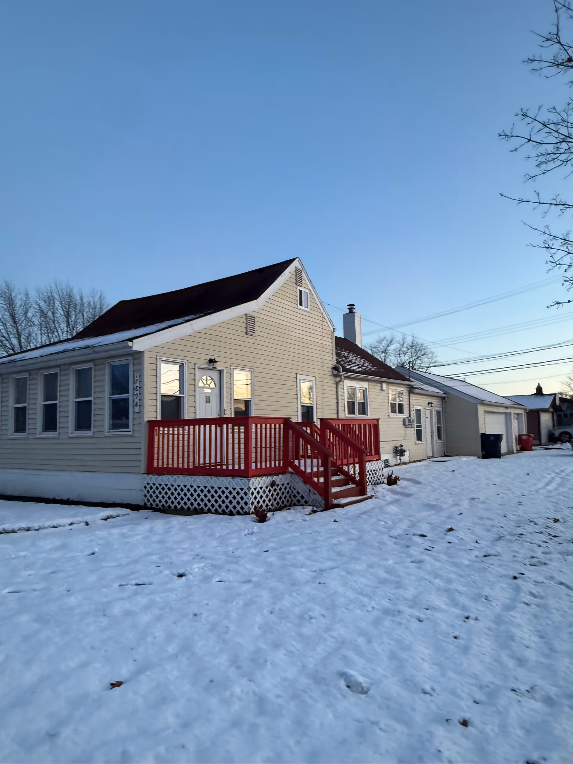 Single-story house with a red railing porch sits in a snow-covered yard under a clear sky.