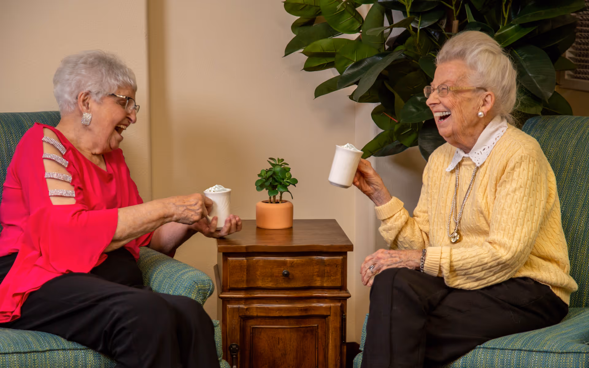 Two elderly women sitting in armchairs facing each other, laughing and holding white mugs with whipped cream. A small wooden side table with a potted plant is between them, and a large green leafy plant is in the background.