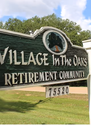 Entrance sign reading 'Village In The Oaks Retirement Community' with address number 75520 on a lawn in front of trees and sky.