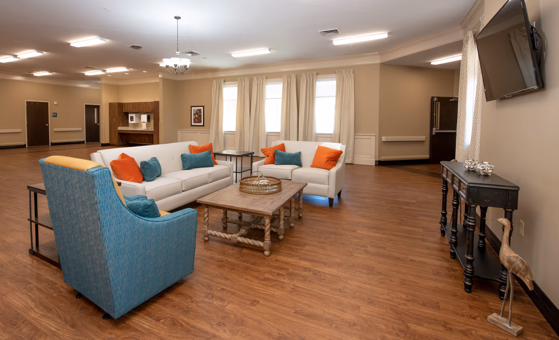 A spacious and well-lit living room area with wooden flooring, featuring two white sofas with orange and teal cushions, a teal armchair, a wooden coffee table, and a black console table with decorative items. The room has beige walls, three windows with light curtains, a wall-mounted TV, and a chandelier hanging from the ceiling.