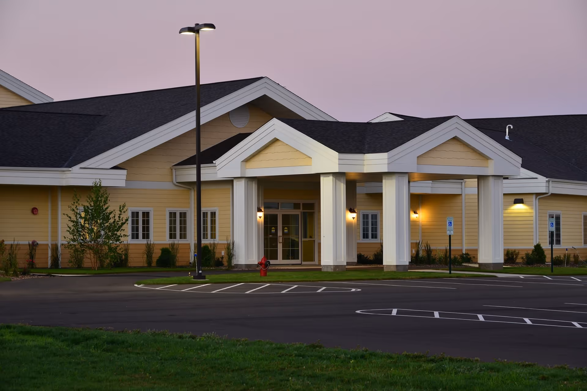 Exterior view of a senior living facility building with a covered entrance supported by white columns, yellow siding, and a dark roof. The parking lot in front has marked spaces including handicapped spots, and a street lamp is lit near the entrance during dusk.