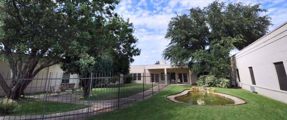 Outdoor courtyard area with a small pond and fountain surrounded by green grass and trees. There is a paved walkway and a metal fence enclosing the space, with beige building walls on either side under a partly cloudy sky.