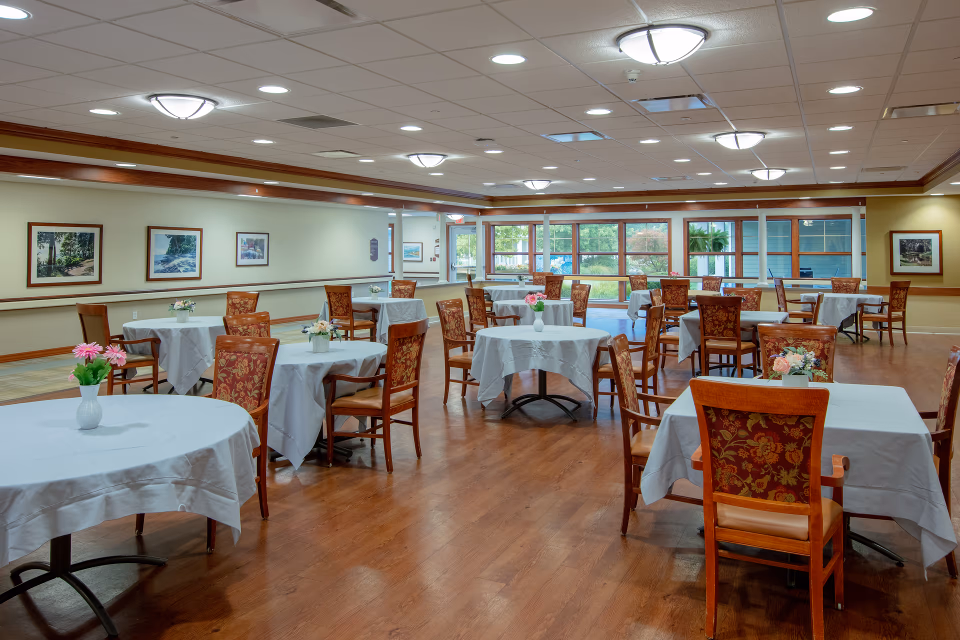 Spacious dining room in a senior living facility with round tables and floral centerpieces.