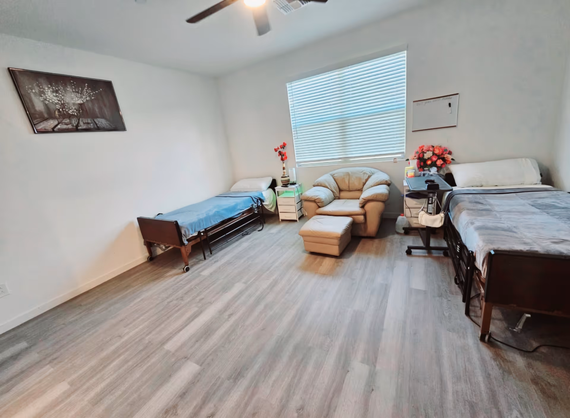 A simple, clean bedroom in an assisted living facility with two single beds, each with a pillow and blanket. Between the beds is a beige armchair with a matching ottoman, positioned in front of a large window with closed blinds. There are small bedside tables next to each bed, decorated with flowers and personal items. The room has light-colored wooden flooring and white walls, with a ceiling fan overhead and a framed picture on one wall.