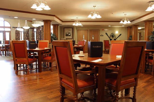A spacious dining room with wooden floors and multiple wooden tables surrounded by red cushioned chairs. Each table has a menu labeled 'Castle Manor' and is set with white napkins and cups. The room is well-lit with multiple ceiling light fixtures and has large windows with patterned curtains allowing natural light to enter.