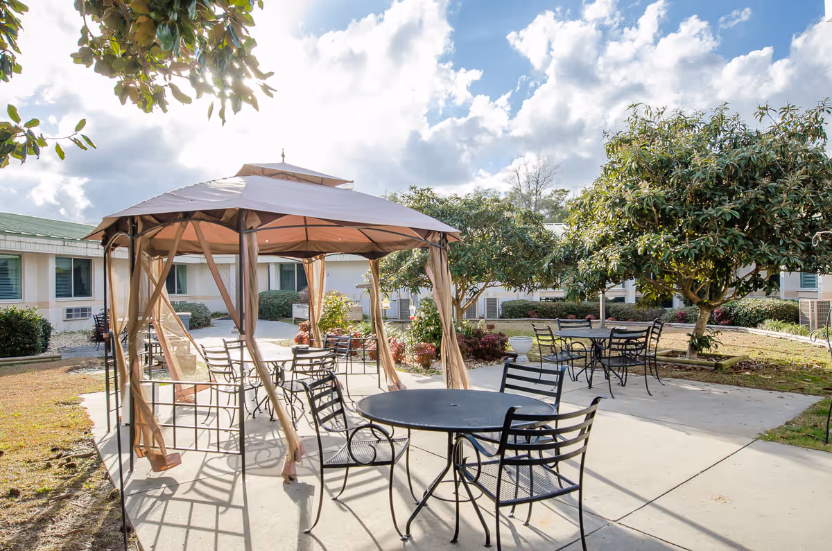 Outdoor patio area at Courtyard Healthcare Center with metal tables and chairs arranged under a gazebo and around the concrete patio, surrounded by trees and bushes with a partly cloudy sky overhead.