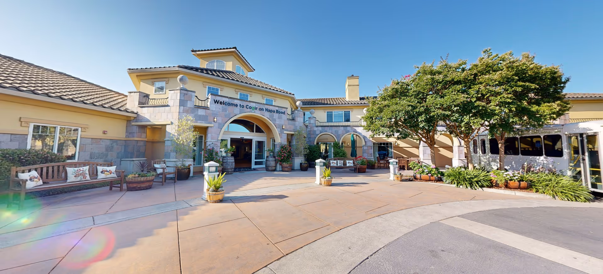 Exterior view of a senior living facility entrance with a curved driveway, benches with cushions, potted plants, and trees under a clear blue sky. The building has a tiled roof and a sign above the entrance that reads 'Welcome to Cogir on Napa Road.'