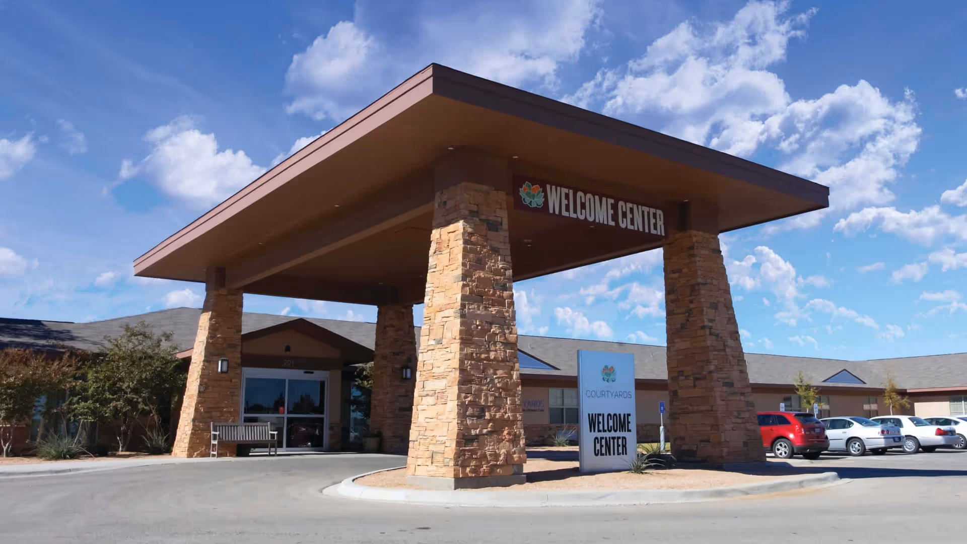 Exterior view of The Courtyards Assisted Living and Memory Care Welcome Center with a large covered entrance supported by stone pillars, a bench near the entrance, and several parked cars under a partly cloudy blue sky.