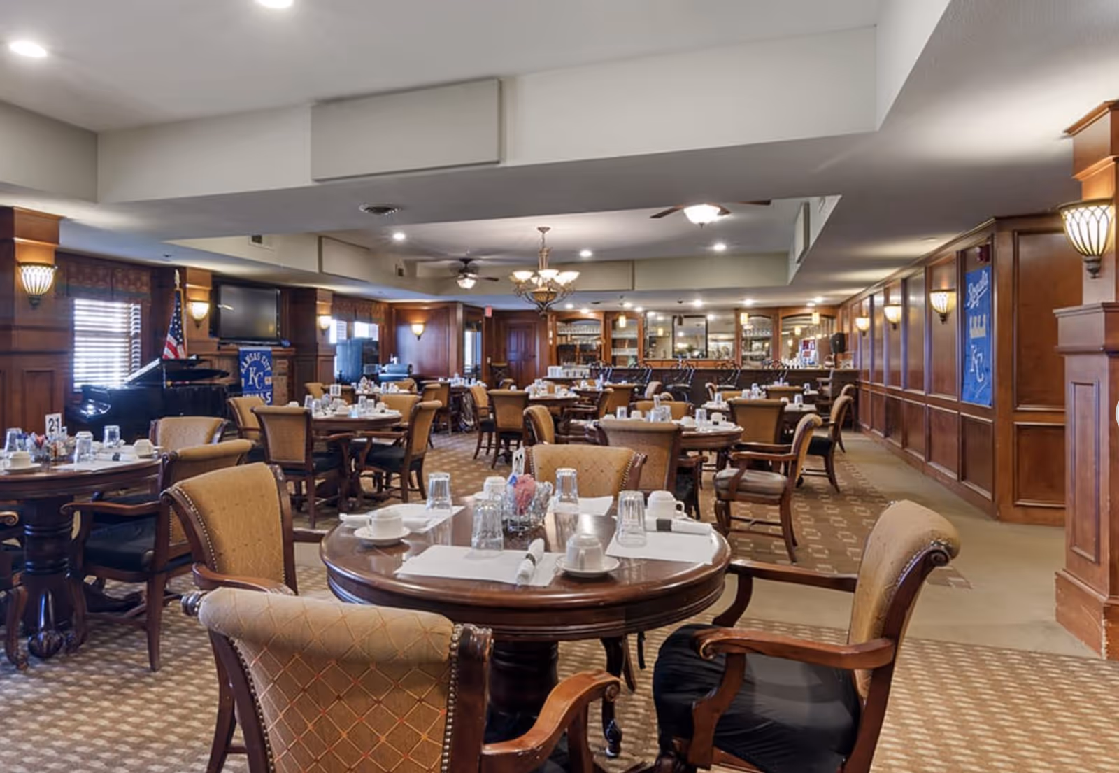 A spacious dining room with multiple round wooden tables and cushioned chairs arranged neatly. Each table is set with white cups, glasses, napkins, and silverware. The room features wood-paneled walls, carpeted floors, ceiling fans, and warm lighting fixtures. An American flag and a piano are visible in the background along with a banner and a television mounted on the wall.