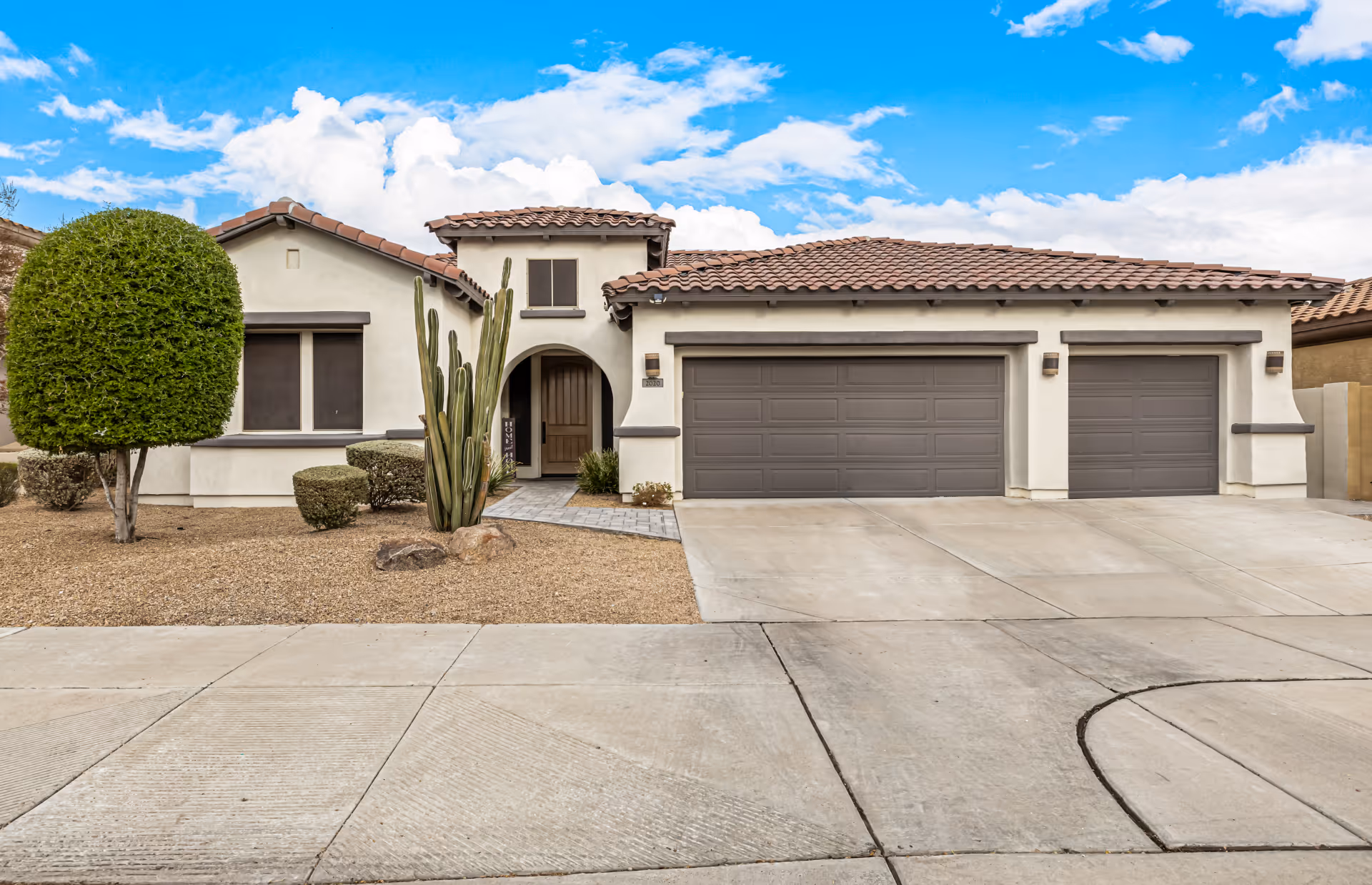 Single-story stucco house with a three-car garage, tiled roof, paved driveway and desert landscaping.