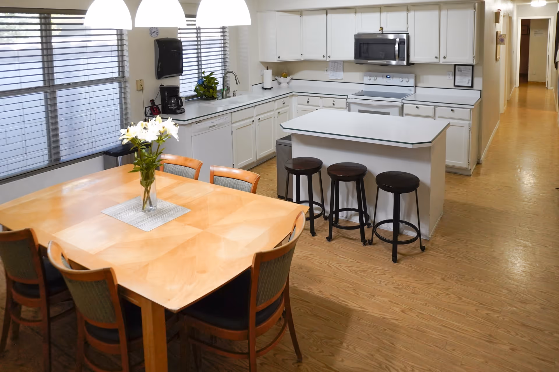 A bright kitchen and dining area with a wooden dining table surrounded by six chairs, a vase with white flowers on the table, white cabinets, a kitchen island with three black stools, a stove, microwave, coffee maker, and large windows with blinds.