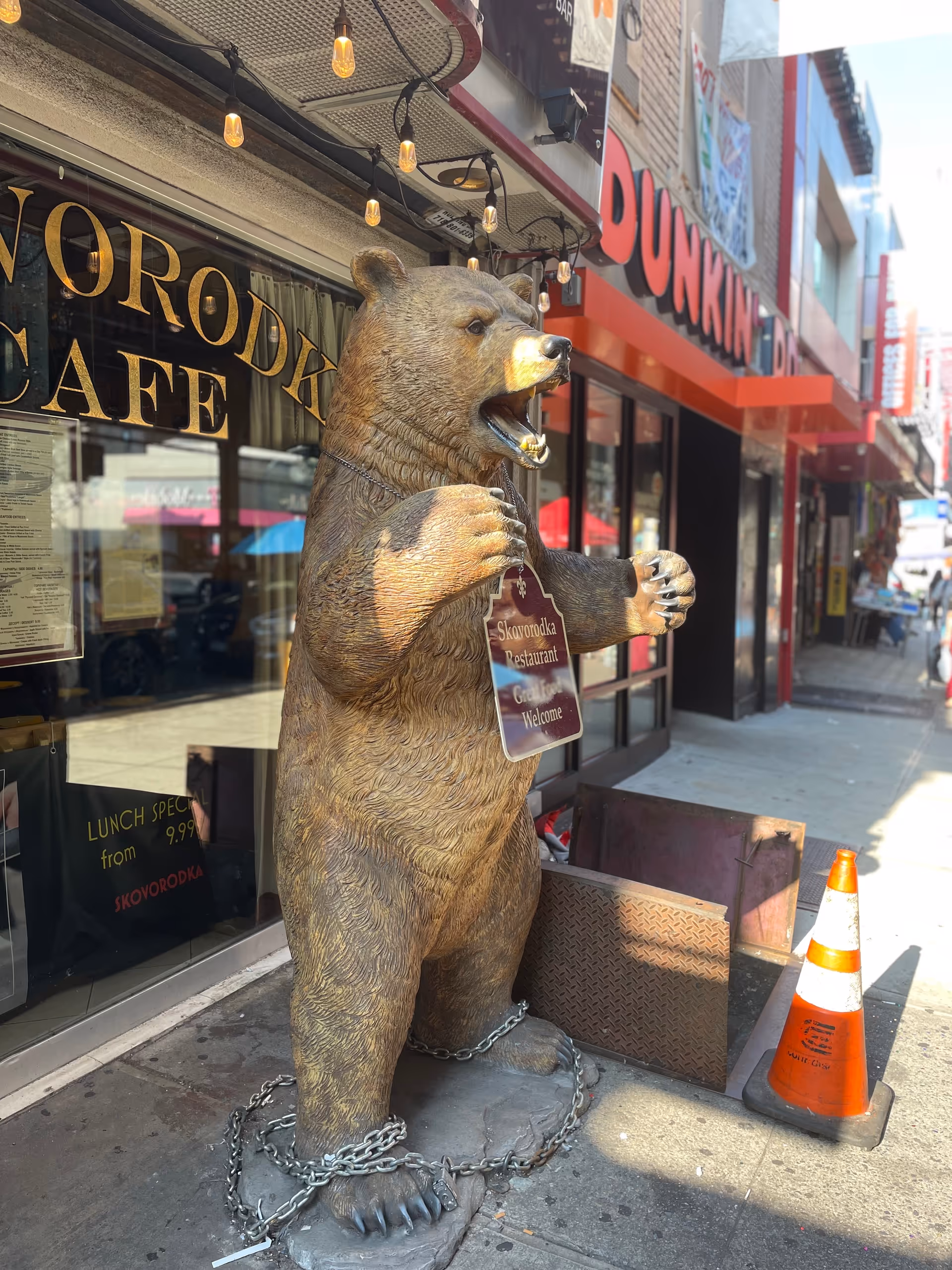 Large bear statue chained to the sidewalk outside a storefront cafe with Dunkin' signage.